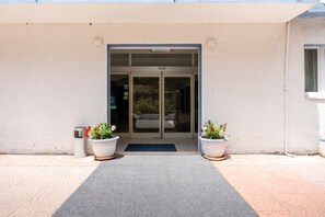 A building entrance with a glass door, potted plants, and a metal trash can. - Hotiday Castelsardo Spiaggia (Lu Bagnu)