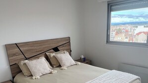 A bedroom with a large bed, a wooden headboard, a nightstand, and a window with a city view. - Sea view apartment (Palavas-les-Flots)