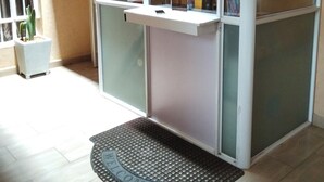 Un espace d’accueil doté d’une porte vitrée, d’un comptoir de réception, d’un paillasson et d’une petite plante.