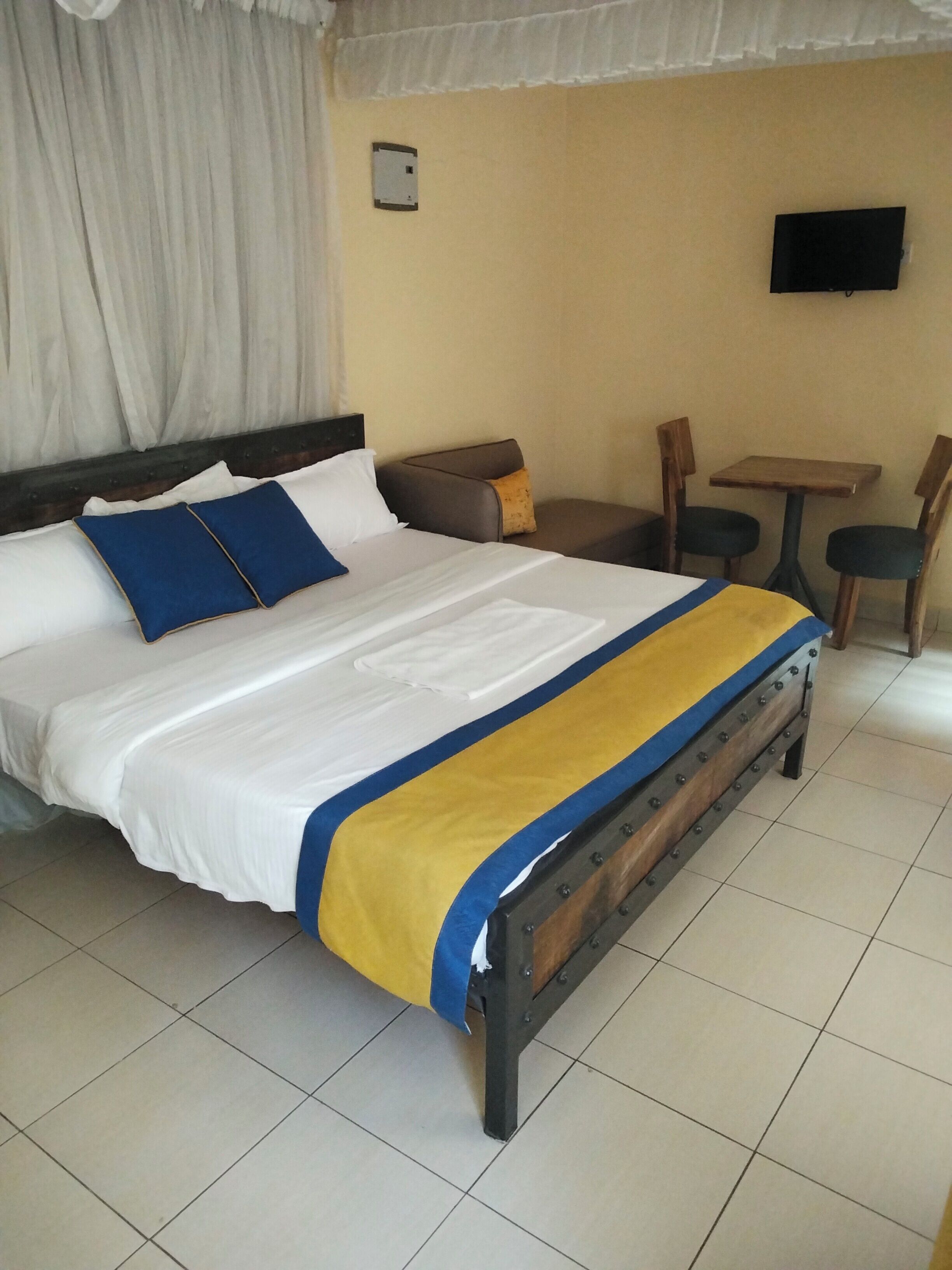 A bed with a white and blue bedspread, a wooden headboard, a small table with chairs, a television on the wall, and a curtain.