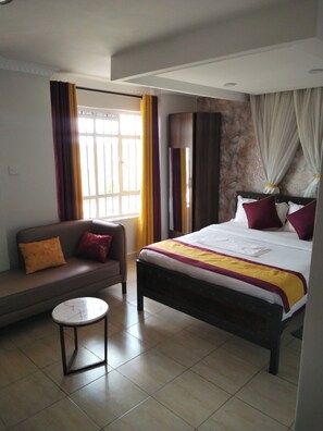 A hotel room with a bed, a sofa, a small table, and a window with curtains. - Heritage plaza accomodation (Nairobi)