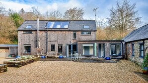 Une maison en bois à deux étages, avec une allée de gravier, une terrasse dotée de mobilier d’extérieur et un bâtiment en pierre attenant.