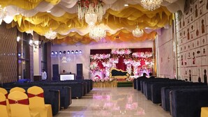 An indoor venue with rows of black chairs, a stage with floral decorations, and chandeliers. - Hotel Awadh Grand (Kanpur)