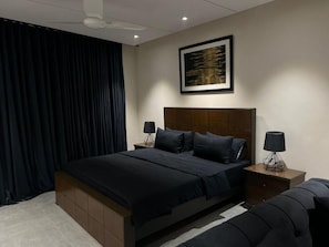 Un dormitorio moderno con una cama grande, cortinas oscuras, una obra de arte enmarcada en la pared y dos mesitas de noche con lámparas.