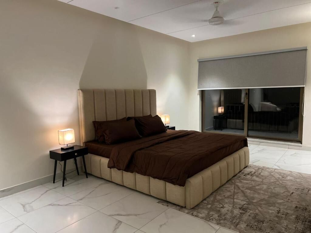 A modern bedroom with a large bed, a nightstand with a lamp, and a view of the outdoors through a large window.