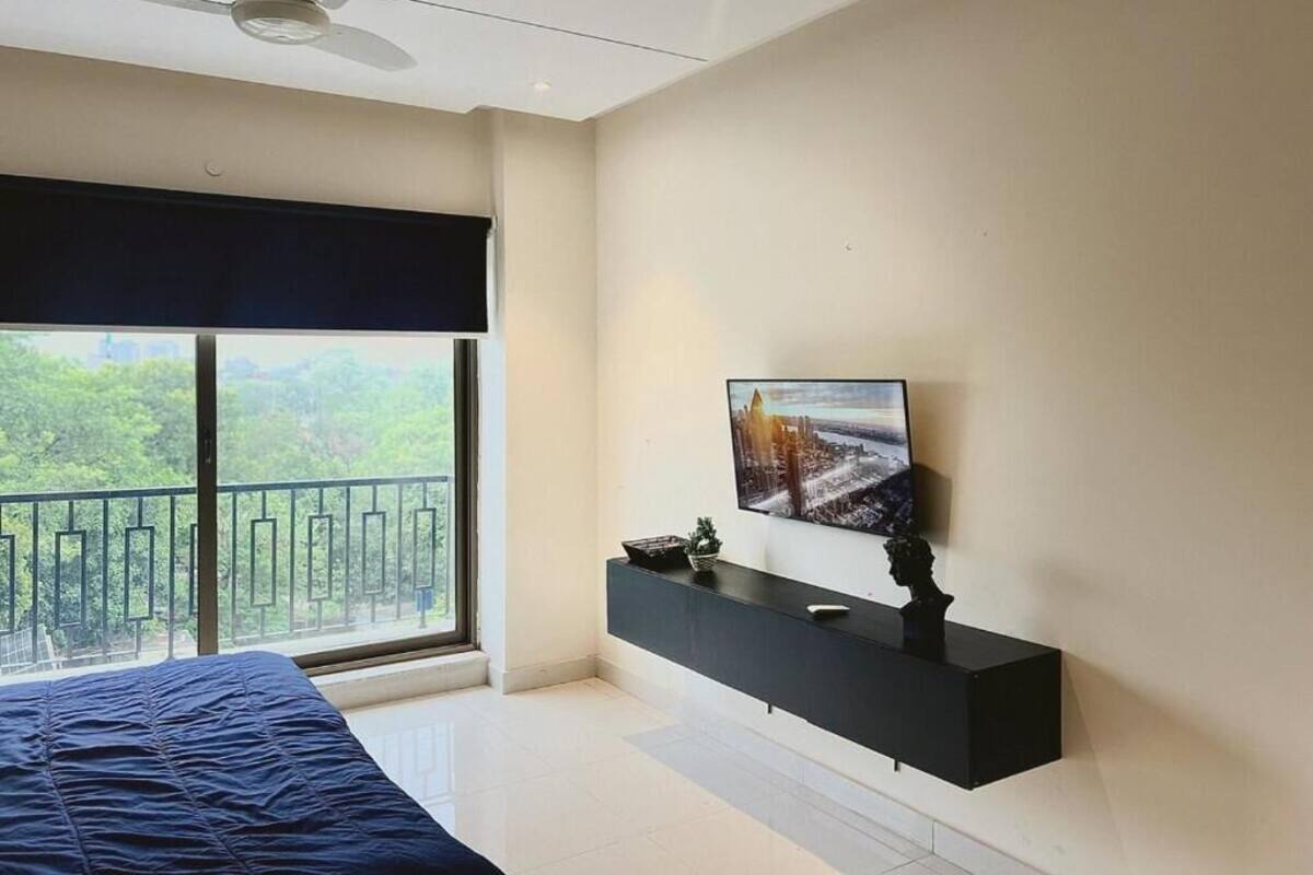 Ein Schlafzimmer mit einem Bett, einem großen Fenster, einem Deckenventilator, einem Fernseher auf einem schwarzen Standfuß und einem Balkon mit Geländer.
