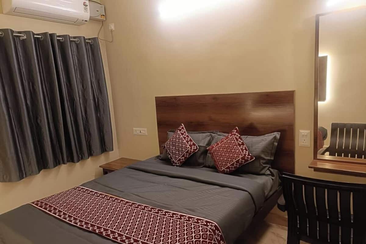 A neatly made bed with a patterned bedspread, two pillows, a wooden headboard, a bedside table, a wall-mounted air conditioner, and a mirror reflecting a chair.