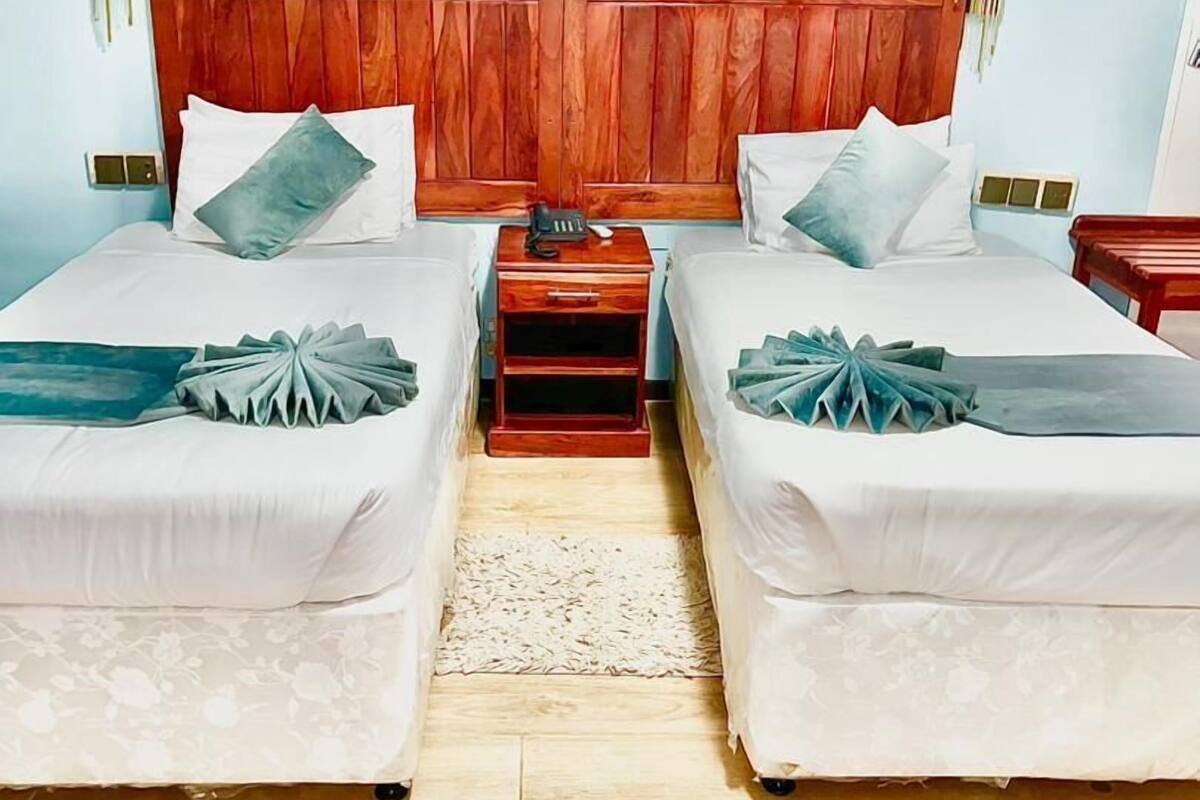 A hotel room with two single beds, a wooden headboard, bedside tables, and a small nightstand with a lamp.