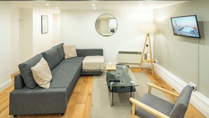 A modern living room with a grey sofa, a glass coffee table, a wooden side table, and a television mounted on the wall. - Stylish Camden 2-Bed Next to Camden Station (London)