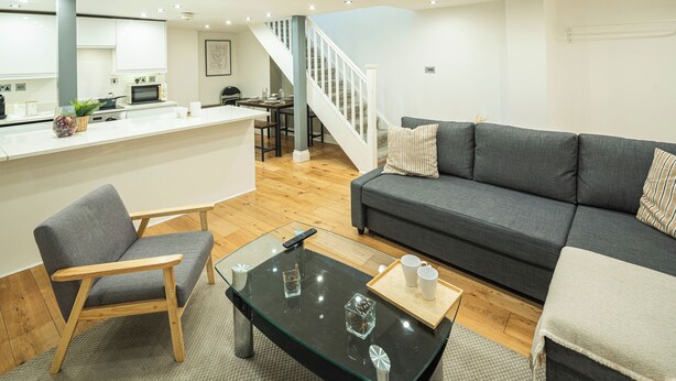 A modern living room with a grey sofa, a wooden chair, a glass coffee table, and a kitchen area in the background. - Stylish Camden 2-Bed Next to Camden Station (London)