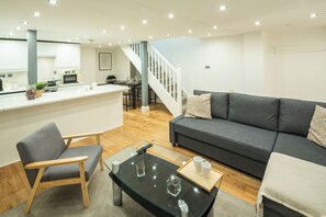 Una sala de estar moderna con un sofá gris, una silla de madera, una mesa de centro de vidrio y una cocina al fondo.