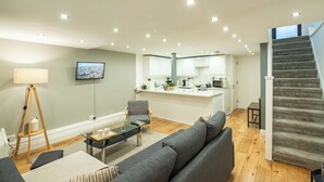 A modern living room with a grey sofa, a glass coffee table, and a lamp. A kitchen with white cabinets and a grey staircase is visible in the background. - Stylish Camden 2-Bed Next to Camden Station (London)