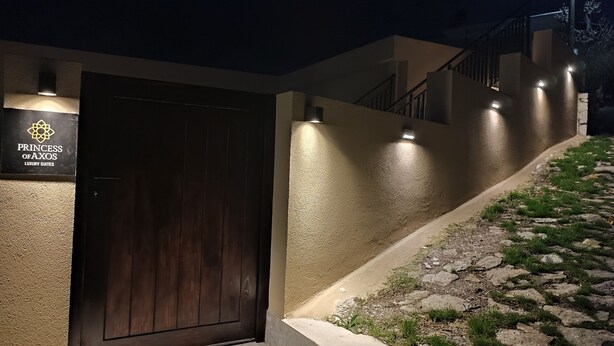 En bygningseingang med en mørk trädør, et skilt der siger "Princess of Axos Luxury Suites", og en stentrappe, der fører op til en oplyst sti.