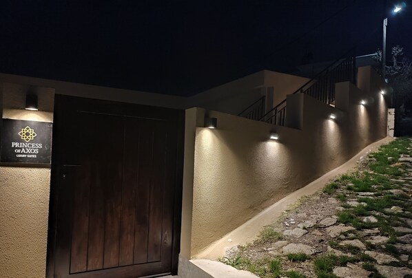 A building entrance with a dark wooden door, a sign reading "Princess of Axos Luxury Suites," and a stone staircase leading up to a lighted pathway. - Axos Getaway: Cozy Junior Apartment for Couples or Solo Travelers (Axos, Mylopotamos)