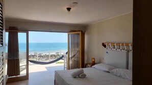 Et hotelværelse med en seng, et natbord, en lampe og udsigt til stranden og havet gennem et stort vindue.
