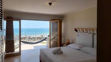 Una habitación de hotel con cama, mesita de noche, lámpara y vistas a la playa y al océano a través de un ventanal grande.