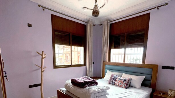 Un lit bien fait, avec une tête de lit, un porte-manteau en bois et une table de chevet avec une lampe.