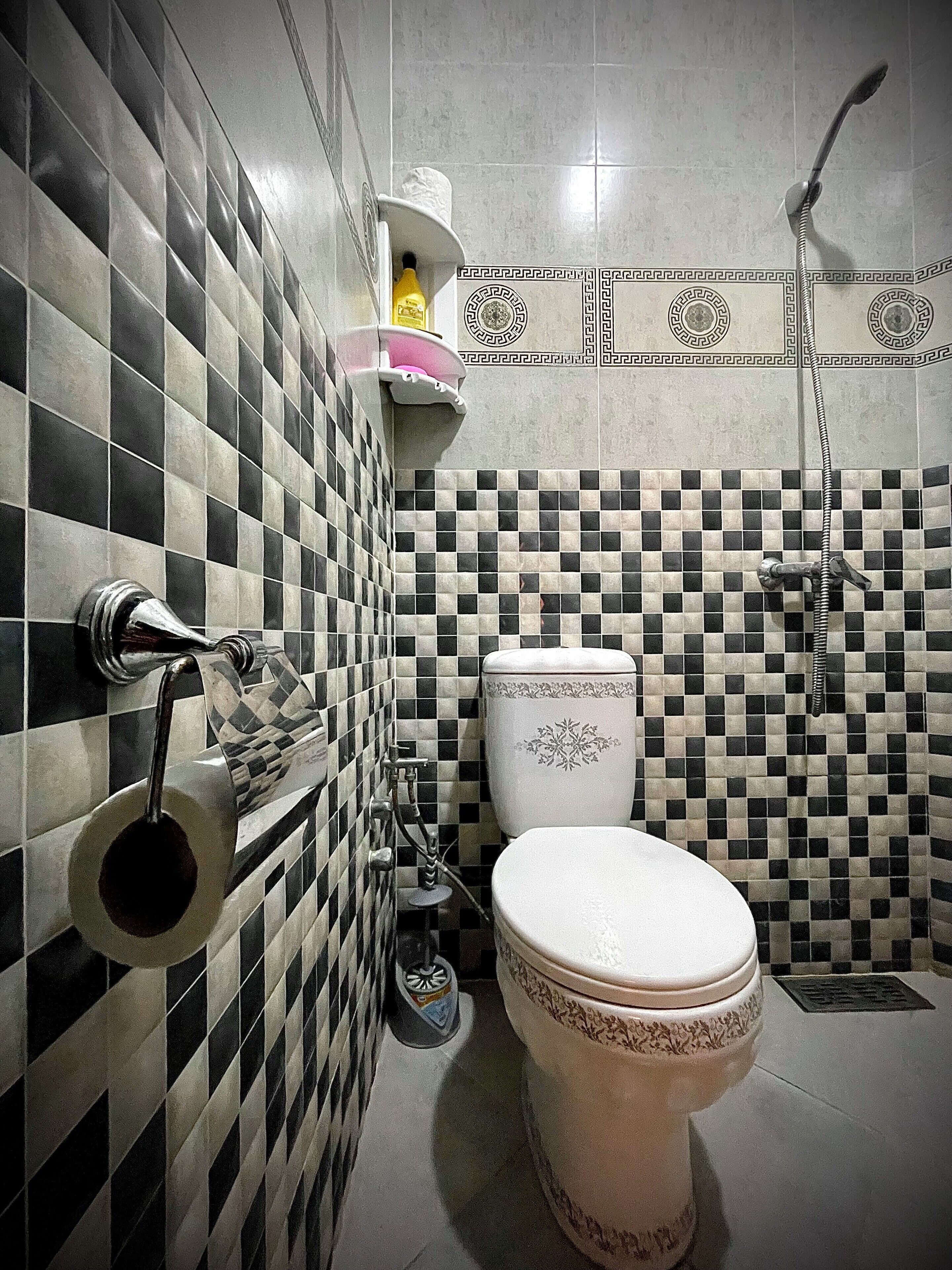 Un baño con suelo de baldosas blancas y negras en damero, un inodoro, una ducha y un dispensador de papel higiénico.