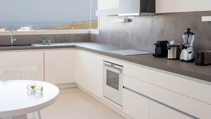 A modern kitchen with a view of the landscape, a white dining table, and a built-in dishwasher. - My Rental Homes - Thera Prince with 2 pools, gym facilitiesand sea view (Fira)