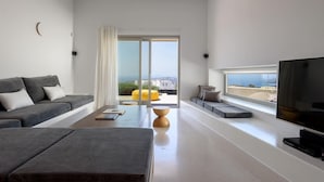 A modern living room with a grey sofa, a coffee table, and a TV. There is a large window with a view of buildings and a yellow chair outside. - My Rental Homes - Thera Prince with 2 pools, gym facilitiesand sea view (Fira)