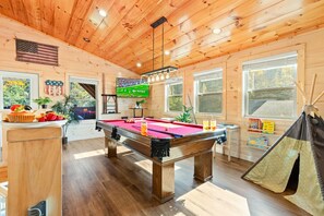 A pool table in a room with wooden walls and ceiling, a window with a view of trees, and a teepee tent. - The Enchanted Cabin by Ghosal Luxury Lodging (Sevierville)