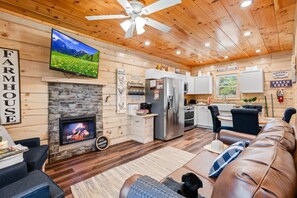 A cozy cabin interior with a stone fireplace, a television mounted above it, a kitchen area with a refrigerator and stove, and a leather couch in the living room. - The Enchanted Cabin by Ghosal Luxury Lodging (Sevierville)