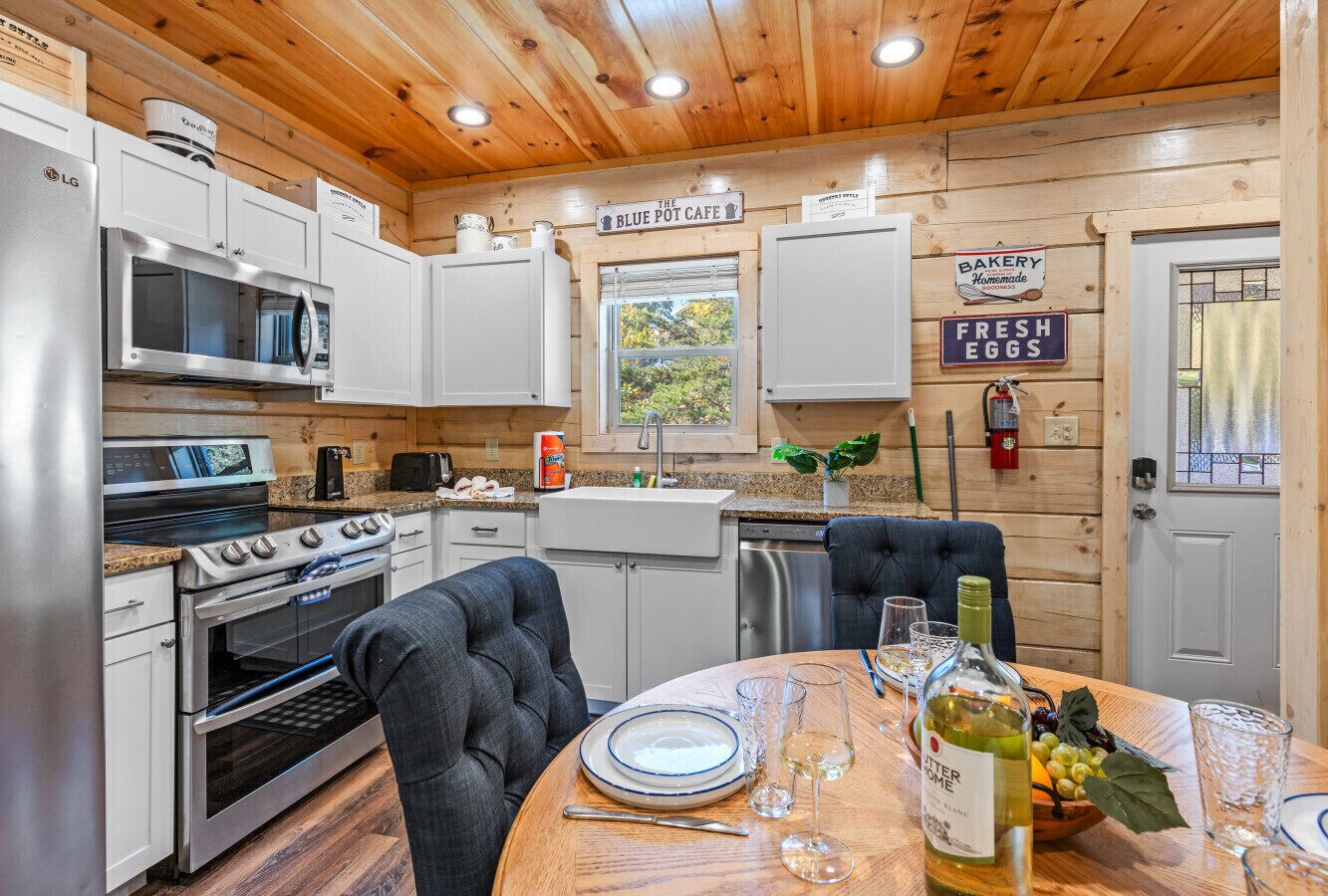 Una cocina con armarios blancos, microondas, horno, una mesa de comedor con platos, vasos y una botella de vino.