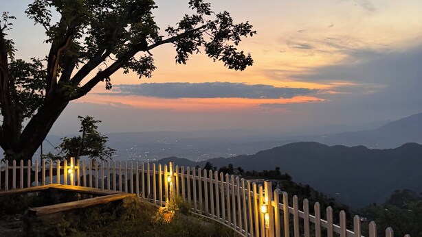 A wooden pathway with a white picket fence, leading to a viewpoint overlooking a sunset over a mountainous landscape. -  Panoramic views of the Doon Valley and Mussoorie Hills - A Serene Hideaway. (Dhaula Giri)