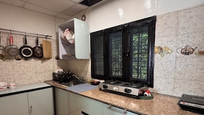 A kitchen with a gas stove, hanging pots, and a window with a view of greenery. -  Panoramic views of the Doon Valley and Mussoorie Hills - A Serene Hideaway. (Dhaula Giri)