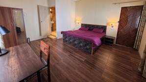A room with a bed, a desk, a chair, a ceiling fan, and a wooden wardrobe. -  Panoramic views of the Doon Valley and Mussoorie Hills - A Serene Hideaway. (Dhaula Giri)