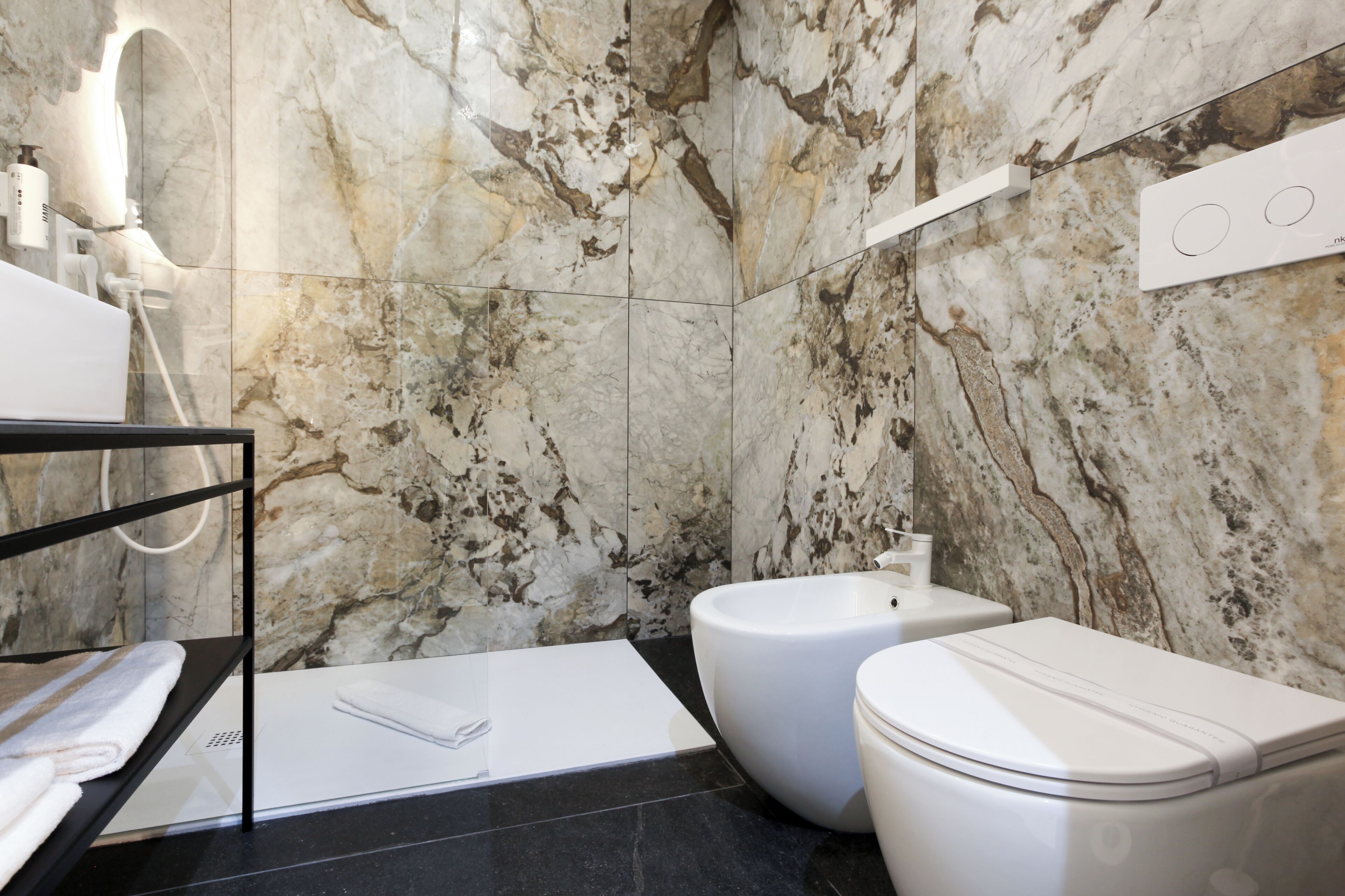 Un bagno moderno con una parete in marmo, un lavandino nero, un WC bianco e un bidet.