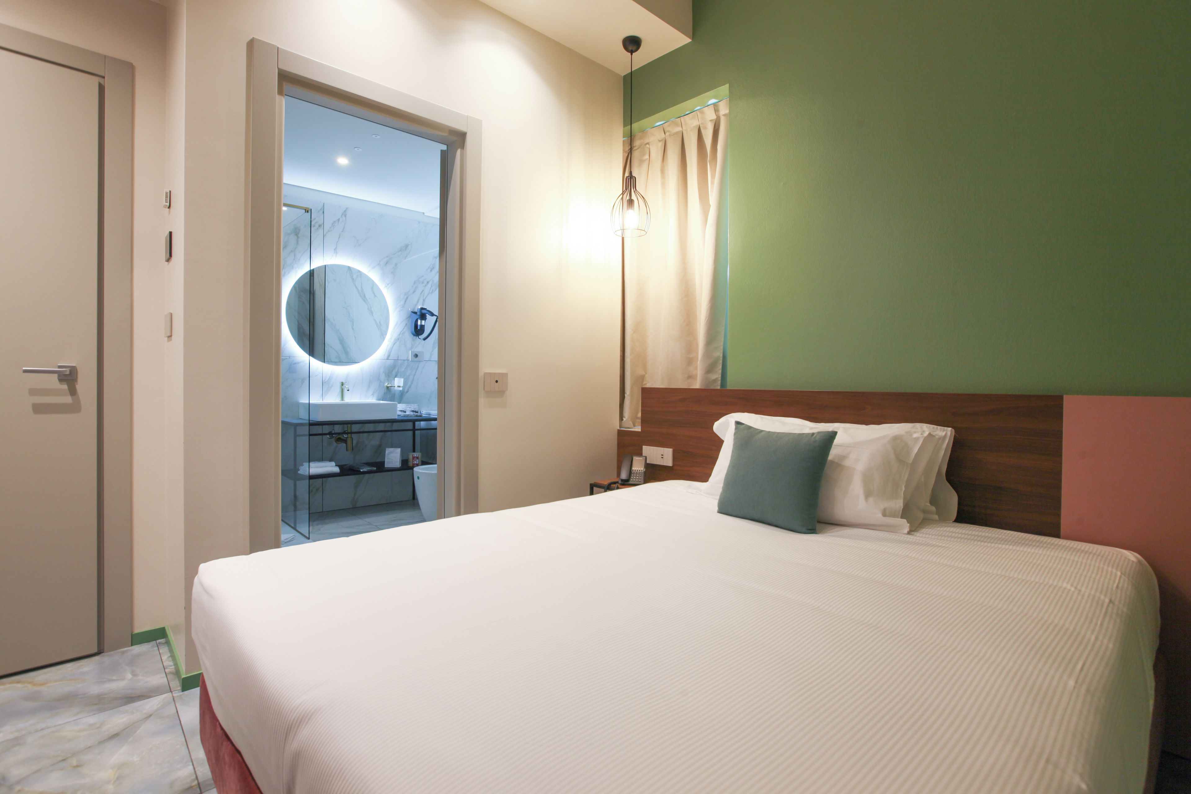 A hotel room with a large bed, a wooden headboard, a bedside table, a mirror with a circular light, and a bathroom visible through an open door.