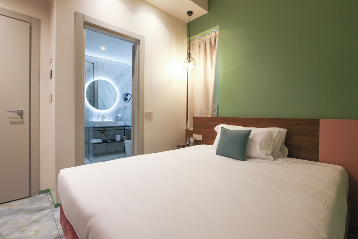A hotel room with a large bed, a wooden headboard, a bedside table, a mirror with a circular light, and a bathroom visible through an open door.