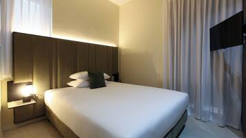 A modern hotel room with a large bed, a bedside table, a wall-mounted TV, and a window with sheer curtains.