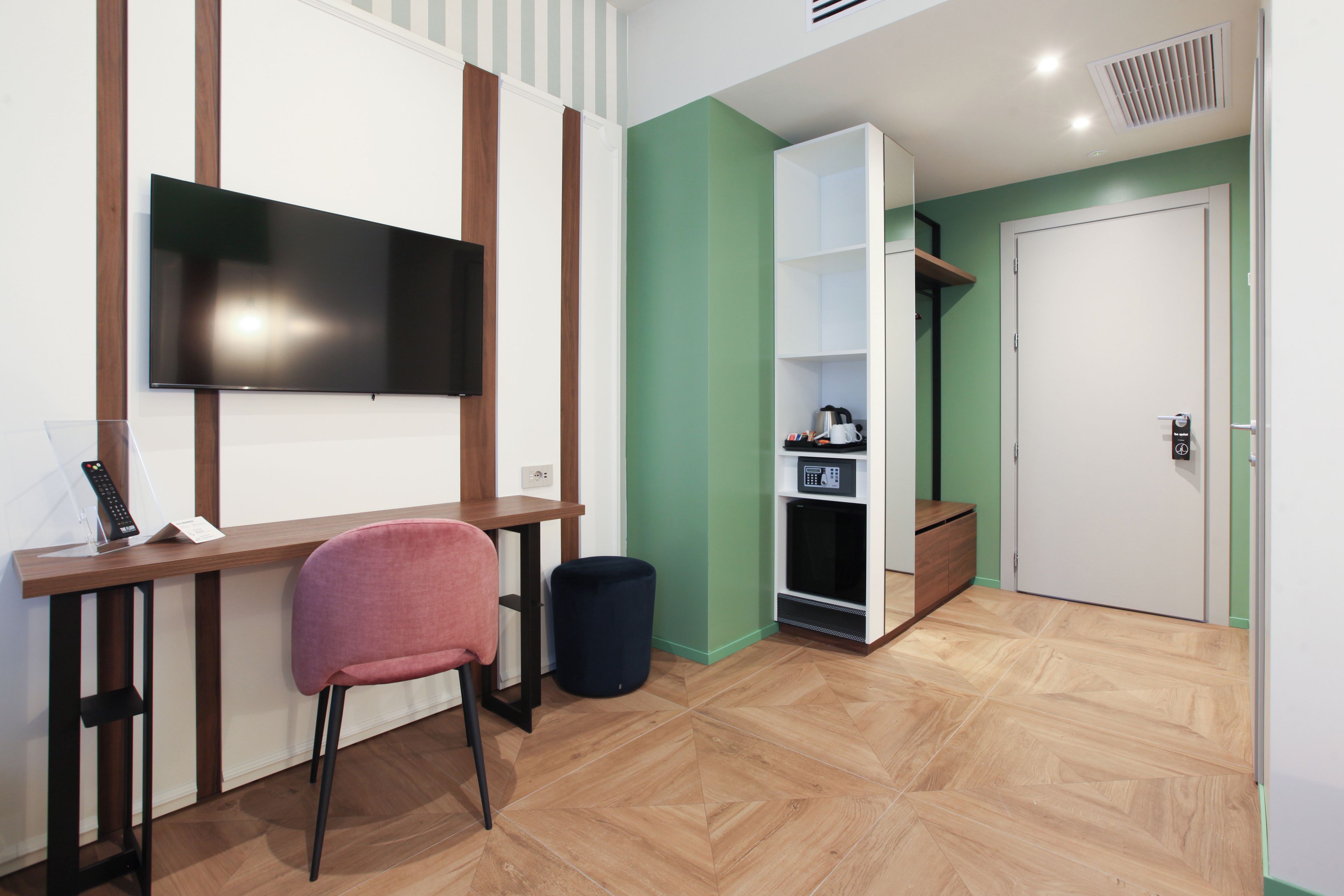A modern room with a flat-screen TV mounted on the wall, a wooden desk with a chair, a small black trash bin, and a built-in shelving unit with a microwave and a coffee maker.