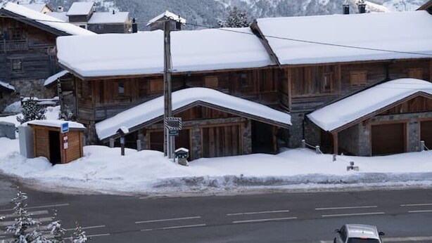 Ein schneebedecktes Holzhaus mit einer Straße im Vordergrund und Bergen im Hintergrund.