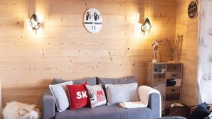 Ein gemütliches Zimmer mit Holzbalkendecke, einem grauen Sofa mit Kissen, einem kleinen Holzschemel, einem Tisch mit gemustertem Tischläufer und einem flauschigen weißen Hund.