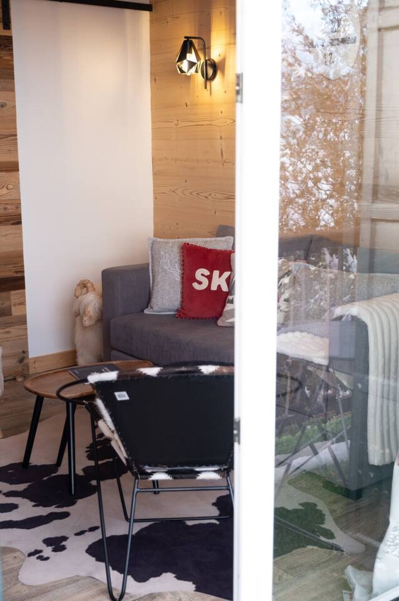 Una habitación acogedora con pared de madera, un sofá gris con un cojín rojo, una silla negra y una pequeña mesa.