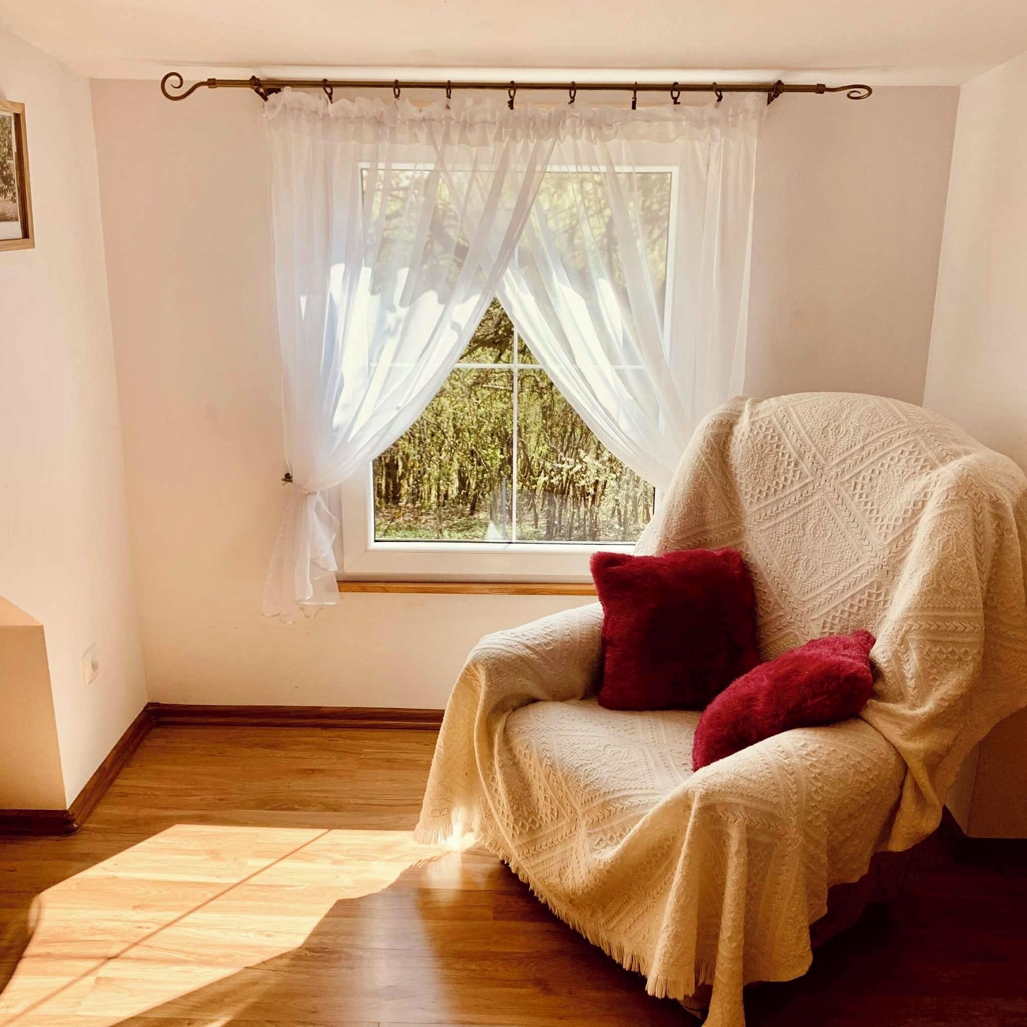 Ein gemütliches Zimmer mit einem großen Fenster, Holzboden und einem weißen Stuhl mit Decke und Kissen.