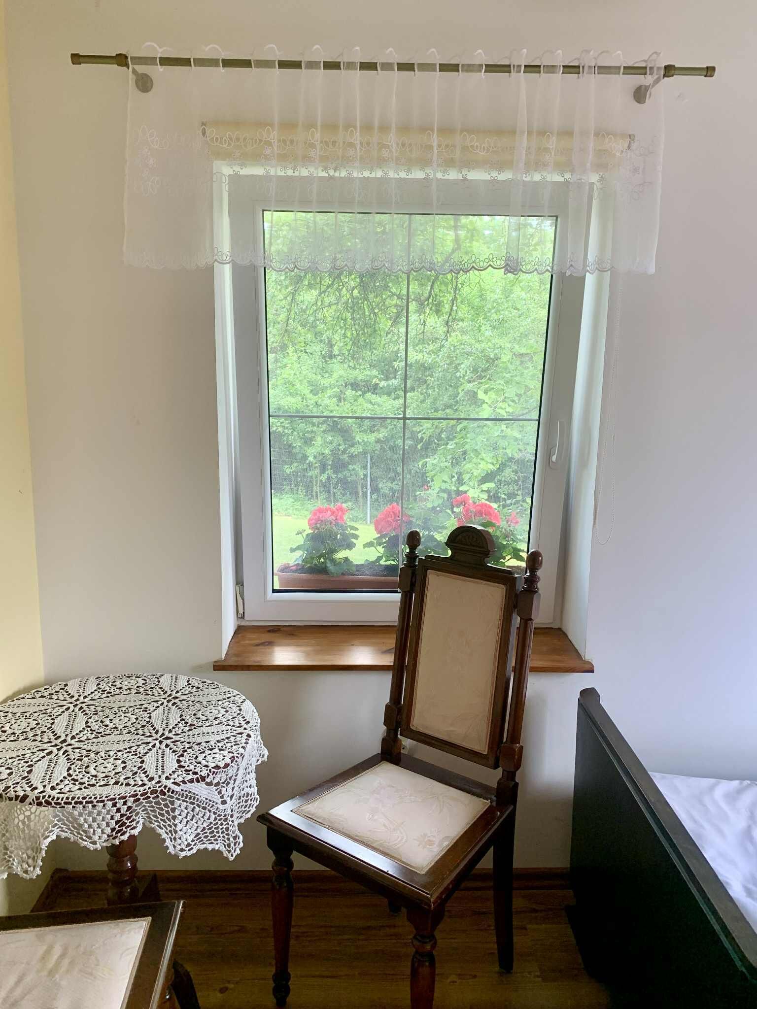 Ein Zimmer mit Holzstuhl an einem Fenster mit Spitzenvorhängen, einem Spitzentisch und einem Bett mit grünem Kopfteil.