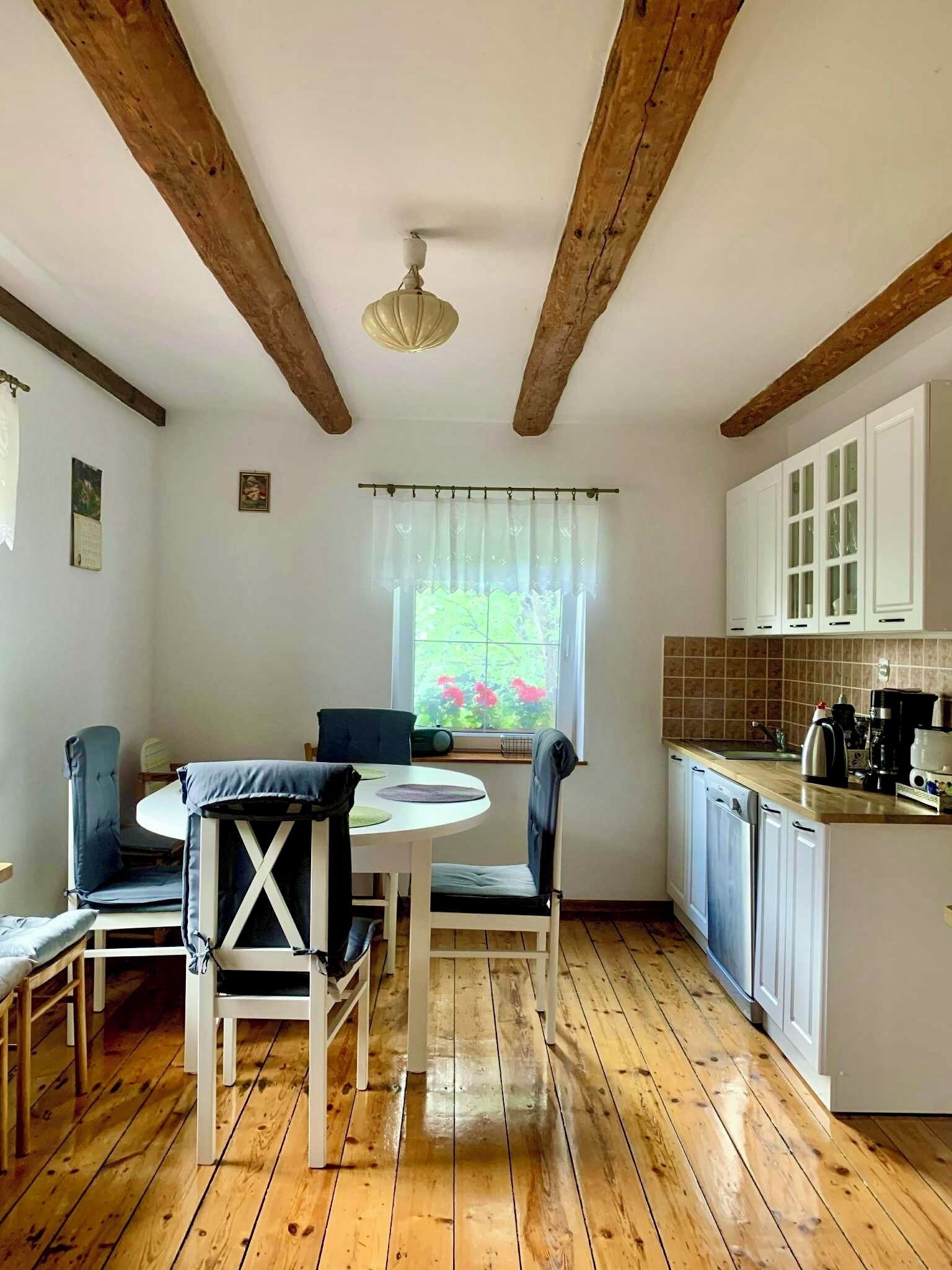 A kitchen with wooden beams, a dining table with chairs, a window with a view of greenery, and white cabinets.