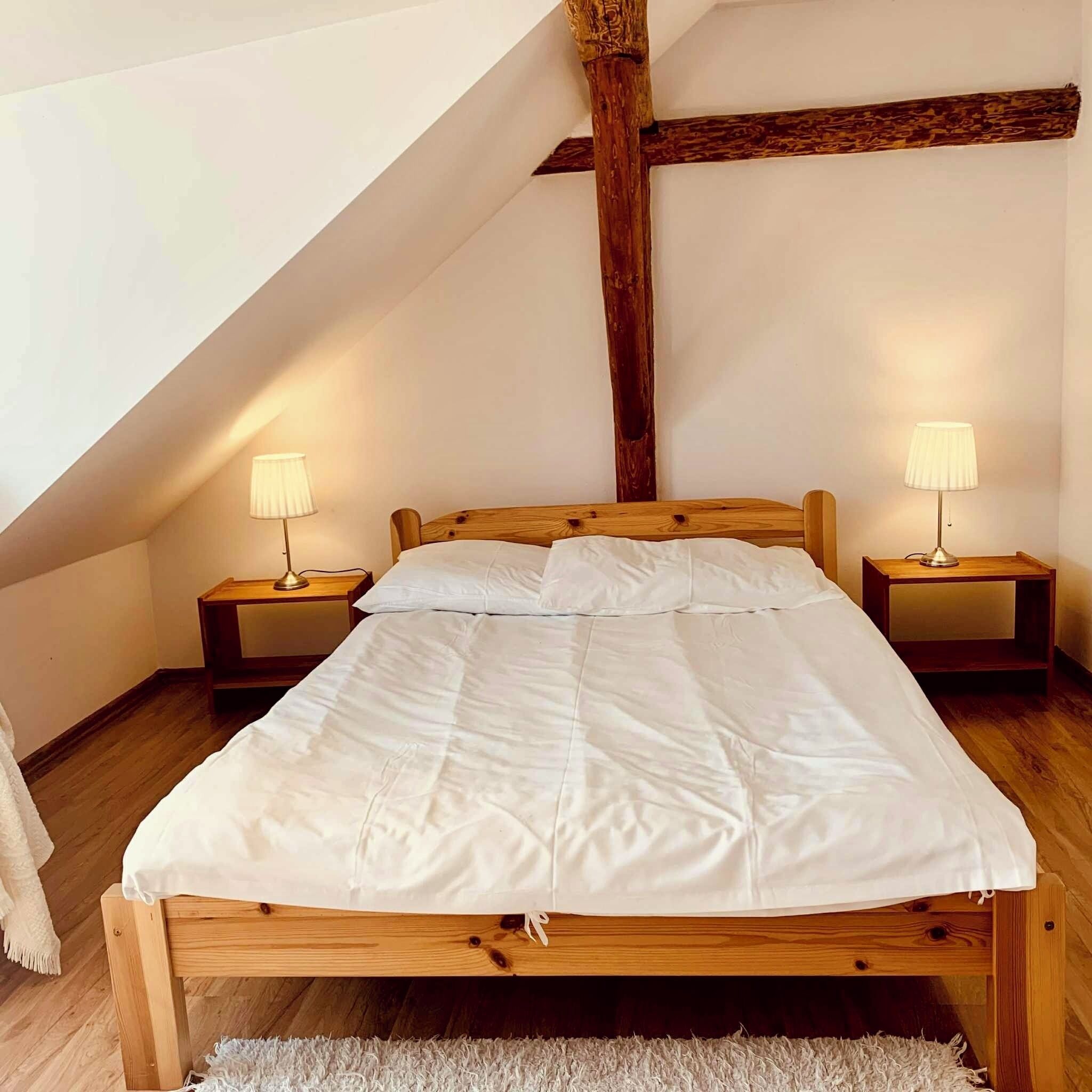 Une chambre à coucher avec un lit en bois, deux tables de chevet avec des lampes, une couvre-lit blanche et un plafond en pente avec des poutres apparentes.