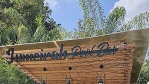 Un espace bar extérieur avec une enseigne en bois indiquant « Jumeirah Pool Bar » et une étagère où sont exposées des bouteilles, ainsi qu’une enseigne pour « The Girls Fort Resort ».