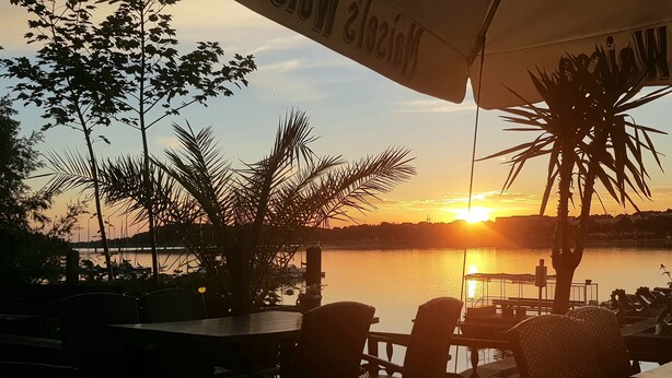 Auringonlaskunäkymä terassilta, jossa on tuoleja ja pöytiä. Terassilta avautuu näkymä tyynelle vesialueelle ja kaukaiseen rantaviivaan.