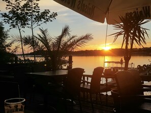 Vista do pôr do sol a partir de um terraço com cadeiras e mesas, com vista para águas calmas e uma linha de costa distante.