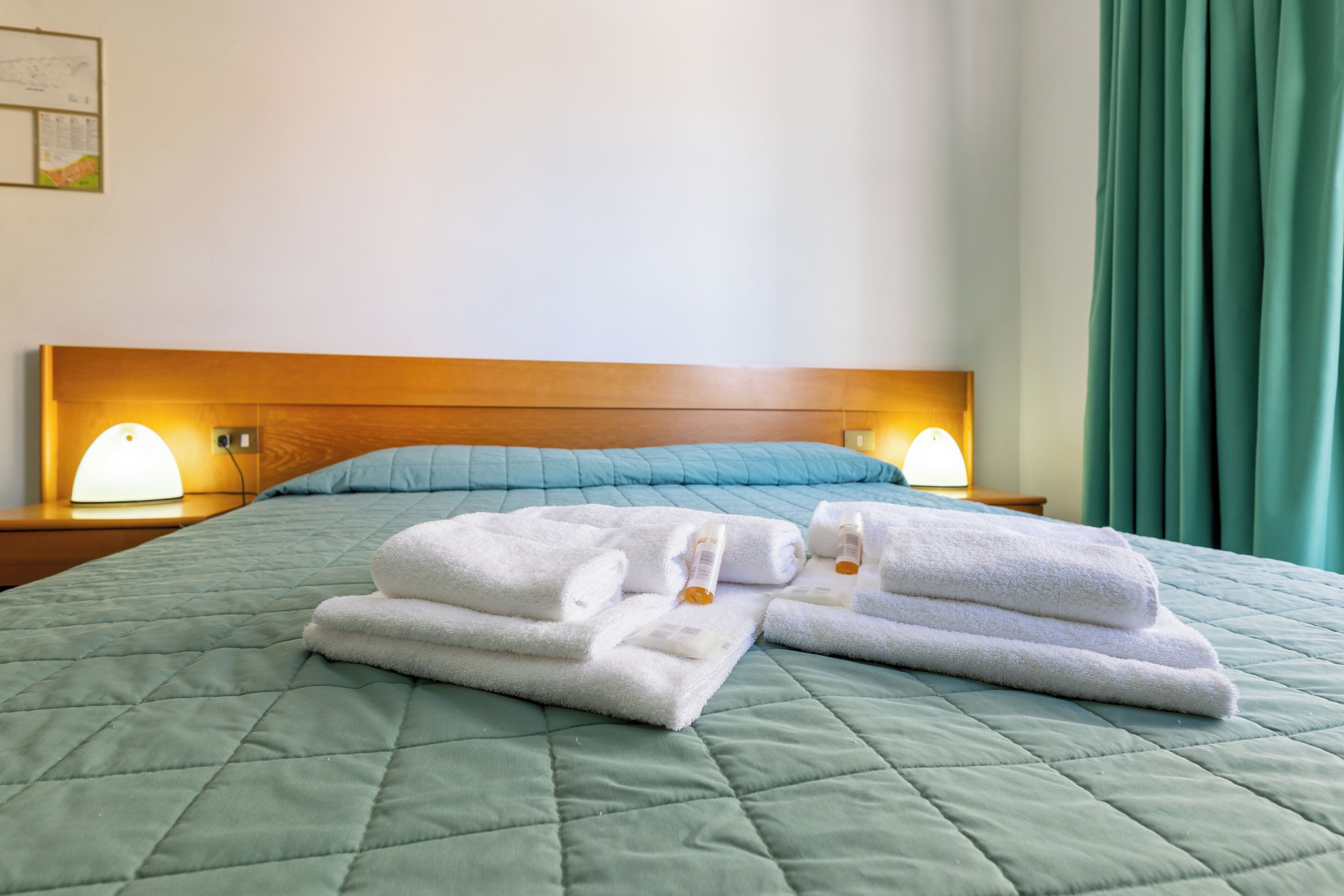 Een opgemaakt bed met een groene gewatteerde spreideken, twee witte handdoeken en twee bedlampjes op een houten hoofdbord.