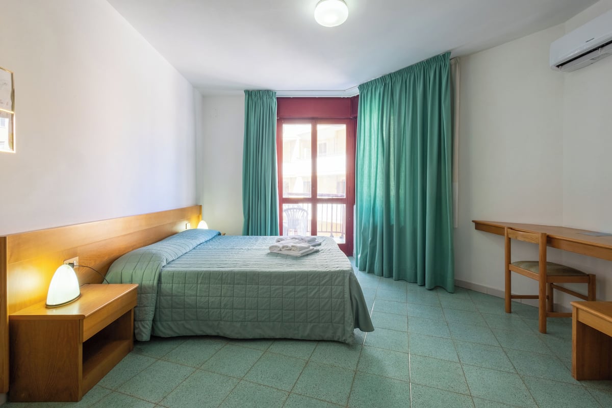 Une chambre à coucher comprenant un lit, une table de chevet, une lampe, un bureau en bois et une fenêtre avec des rideaux.