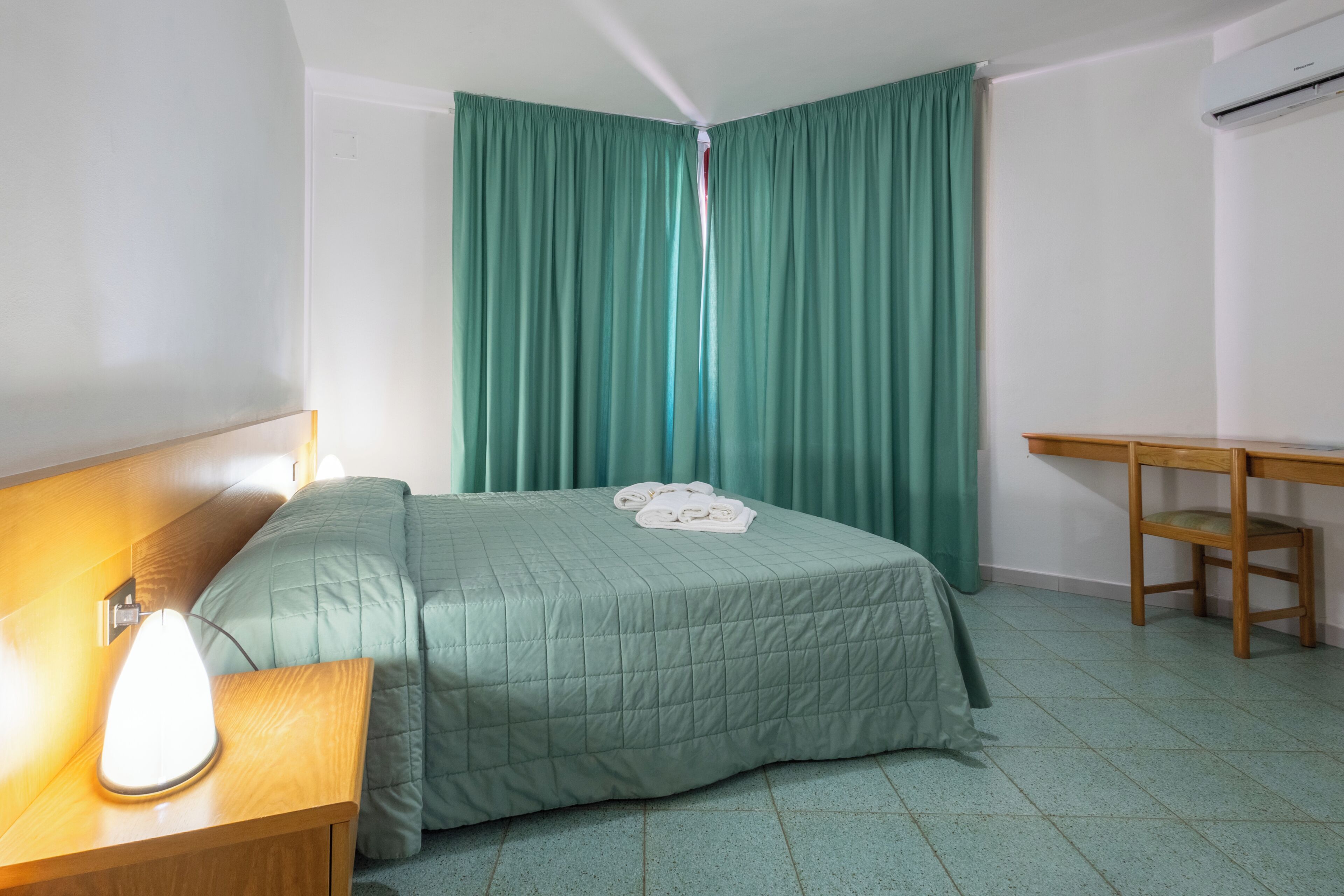 A room with a bed, a nightstand with a lamp, a wooden desk, and a window with green curtains.