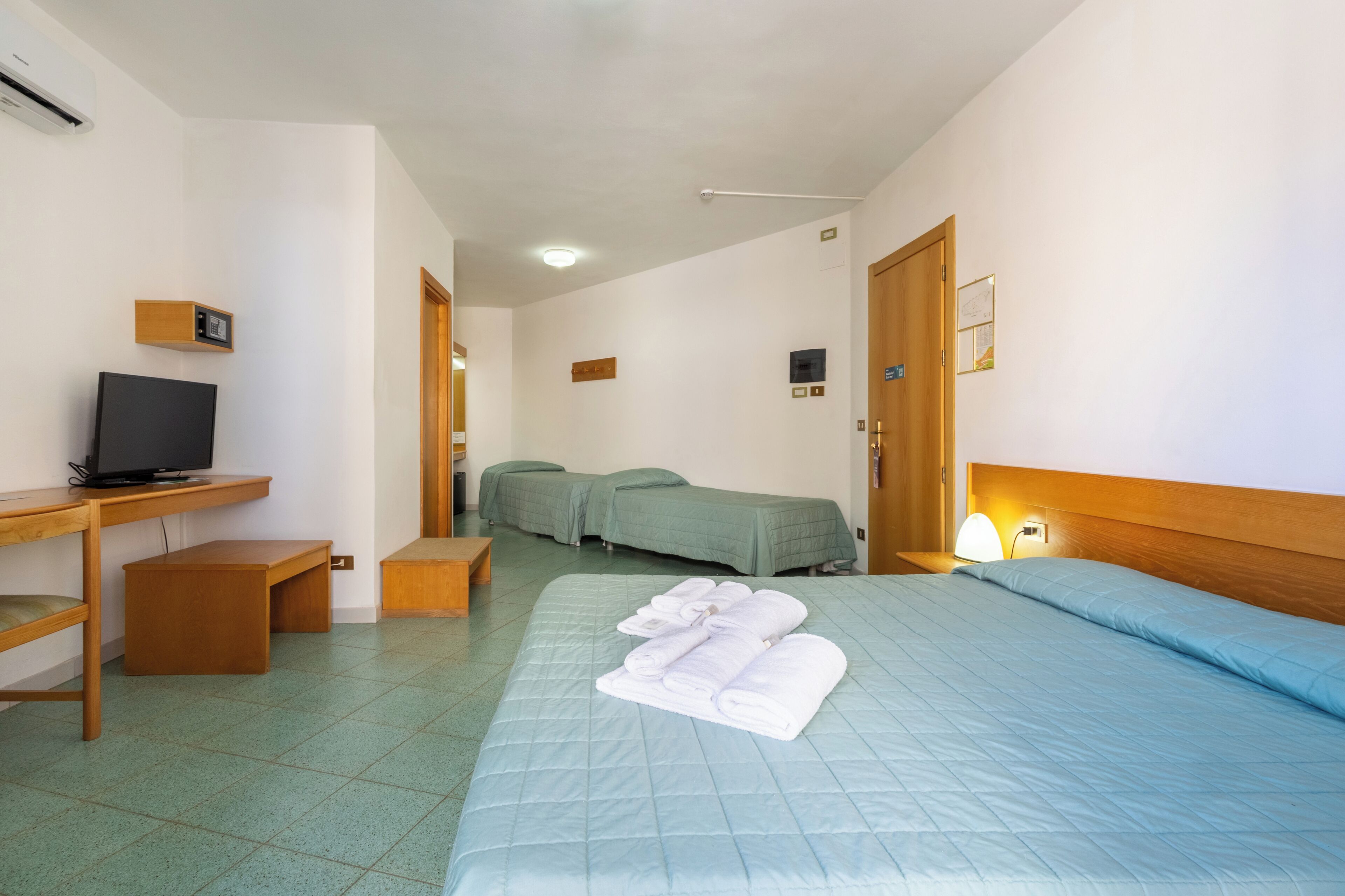 A hotel room with a bed, a desk with a television, a bedside lamp, and a small wooden bench.