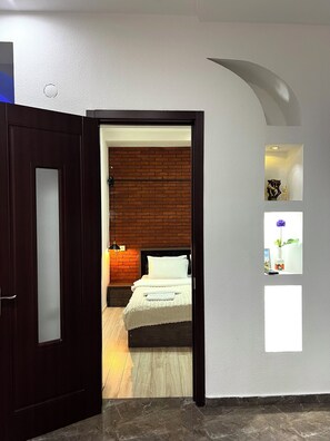 Une pièce avec un lit, un mur en briques, un plancher en bois, une porte sombre et un mur blanc avec une petite étagère.